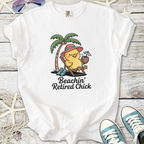 White t-shirt featuring a cartoon chick relaxing under a palm tree with drink, text 'Beachin' Retired Chick'.