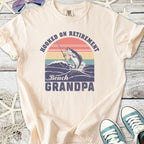 Cream t-shirt with 'Hooked on Retirement Beach Grandpa' graphic featuring a fishing theme and colorful sunset design.