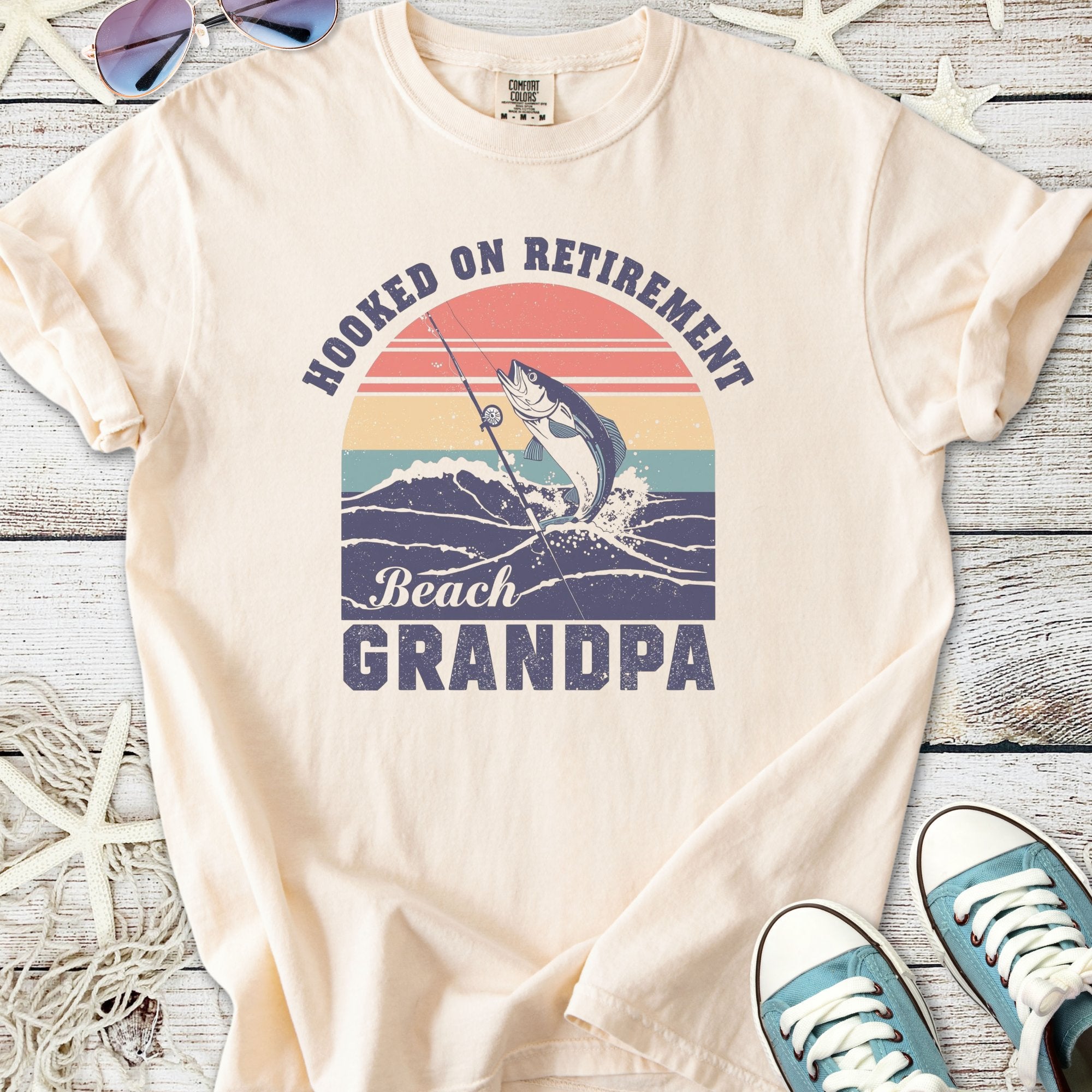 Cream t-shirt with 'Hooked on Retirement Beach Grandpa' graphic featuring a fishing theme and colorful sunset design.