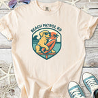 Light beige t-shirt featuring a Beach Patrol K9 graphic with a dog, surfboard, and palm tree.