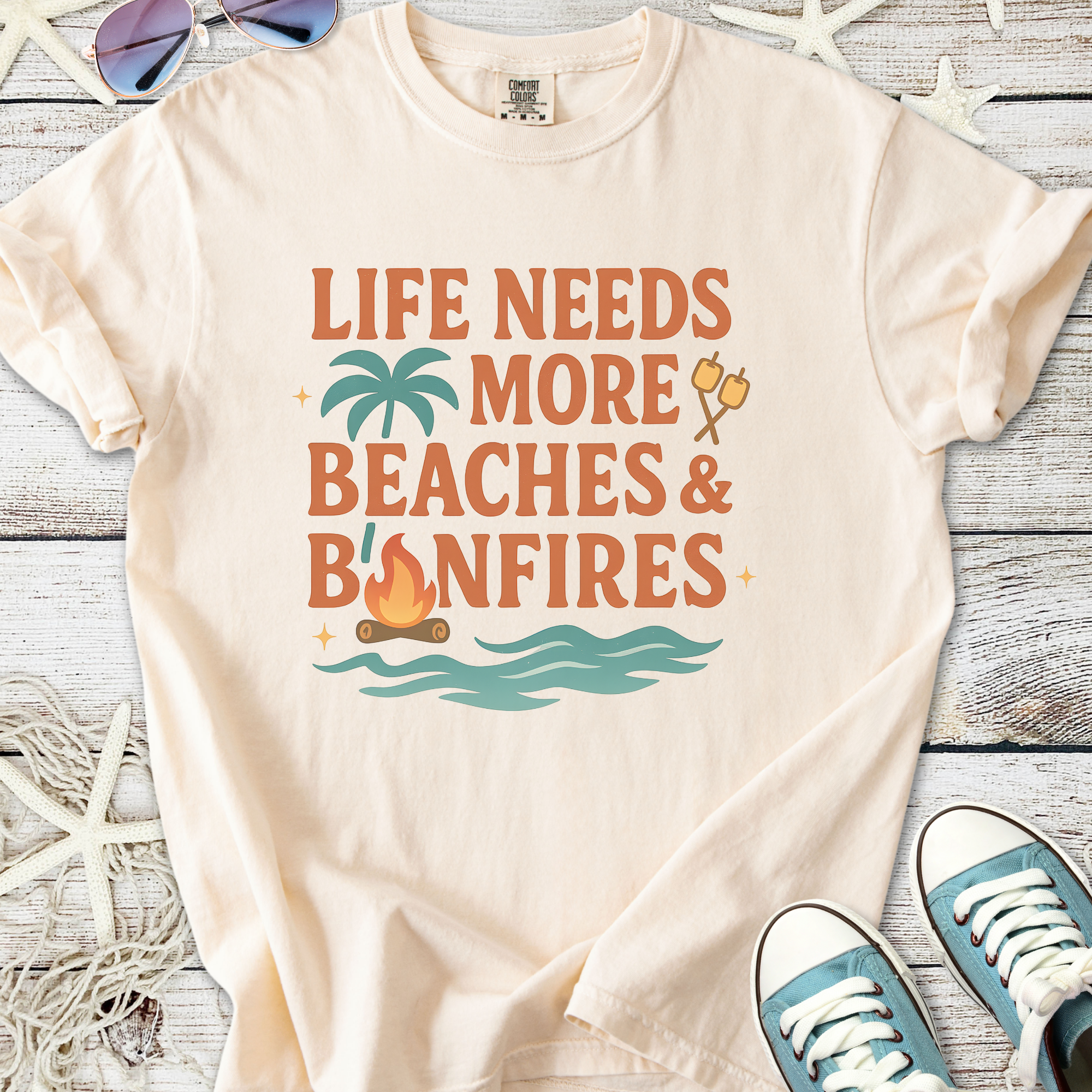 Light beige t-shirt featuring 'Life Needs More Beaches & Bonfires' graphic with palm trees and campfire.