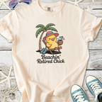 Peach-colored t-shirt featuring a cute chick under a palm tree, sipping a drink, labeled 'Beachin’ Retired Chick'.