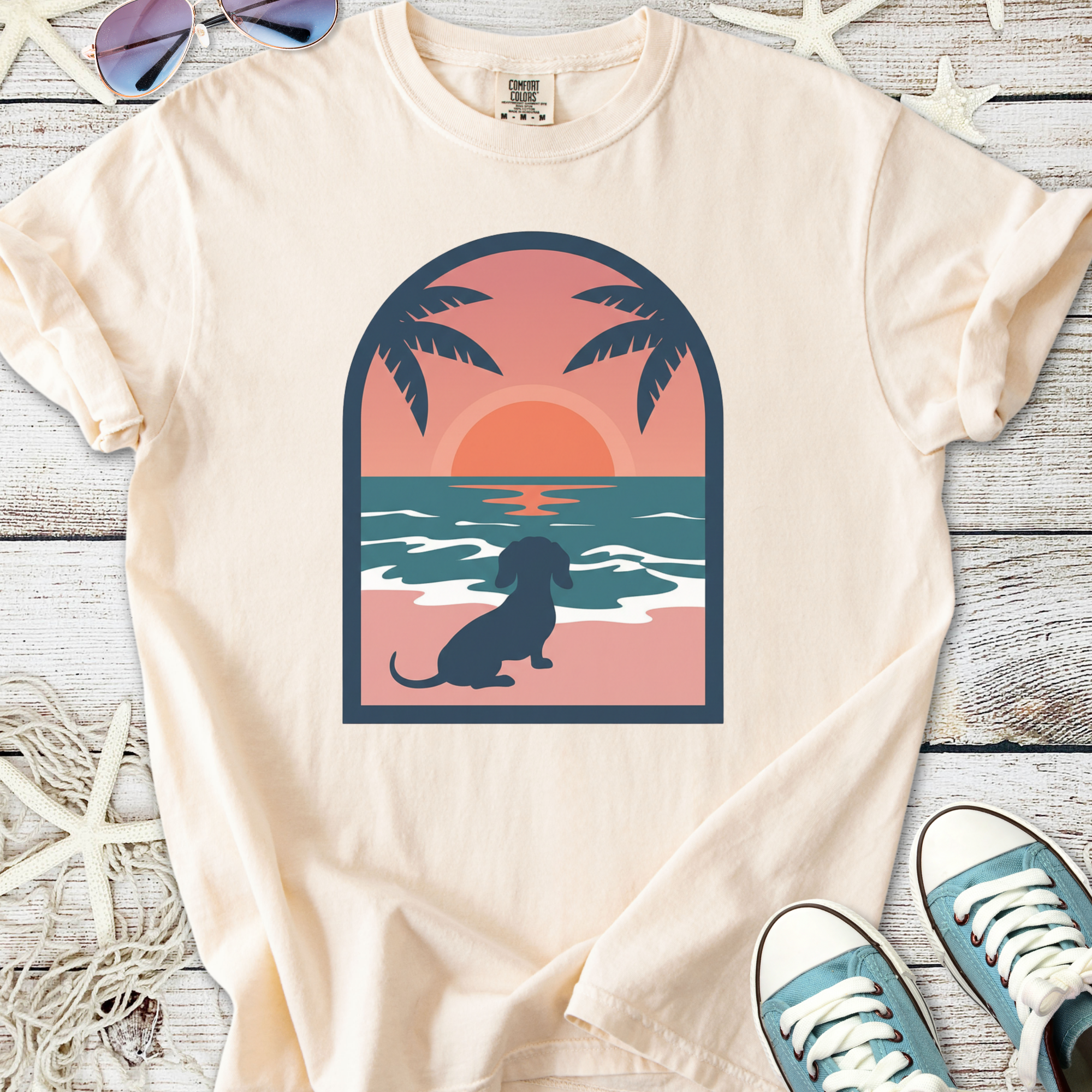 Cream t-shirt featuring a dachshund silhouette against a sunset beach scene with palm trees.