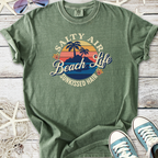 Green cotton t-shirt featuring 'Salty Air Beach Life' graphic with palm trees and sunset design.