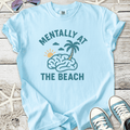 Light blue t-shirt with 'Mentally at the Beach' graphic, featuring a brain, palm tree, and sun.