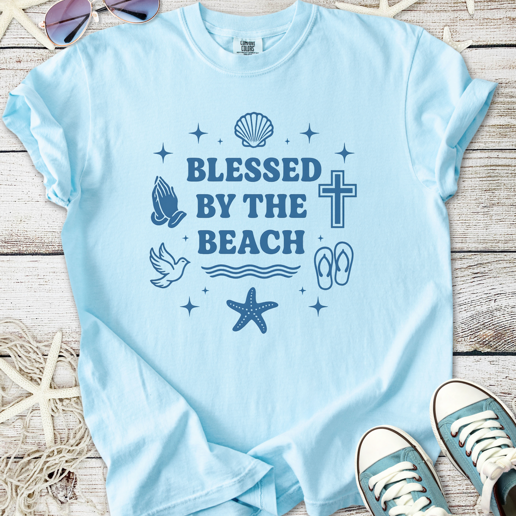 Light blue t-shirt with 'Blessed by the Beach' graphic featuring seashells, a cross, and beach motifs.