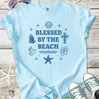 Light blue t-shirt with 'Blessed by the Beach' graphic featuring seashells, a cross, and beach motifs.
