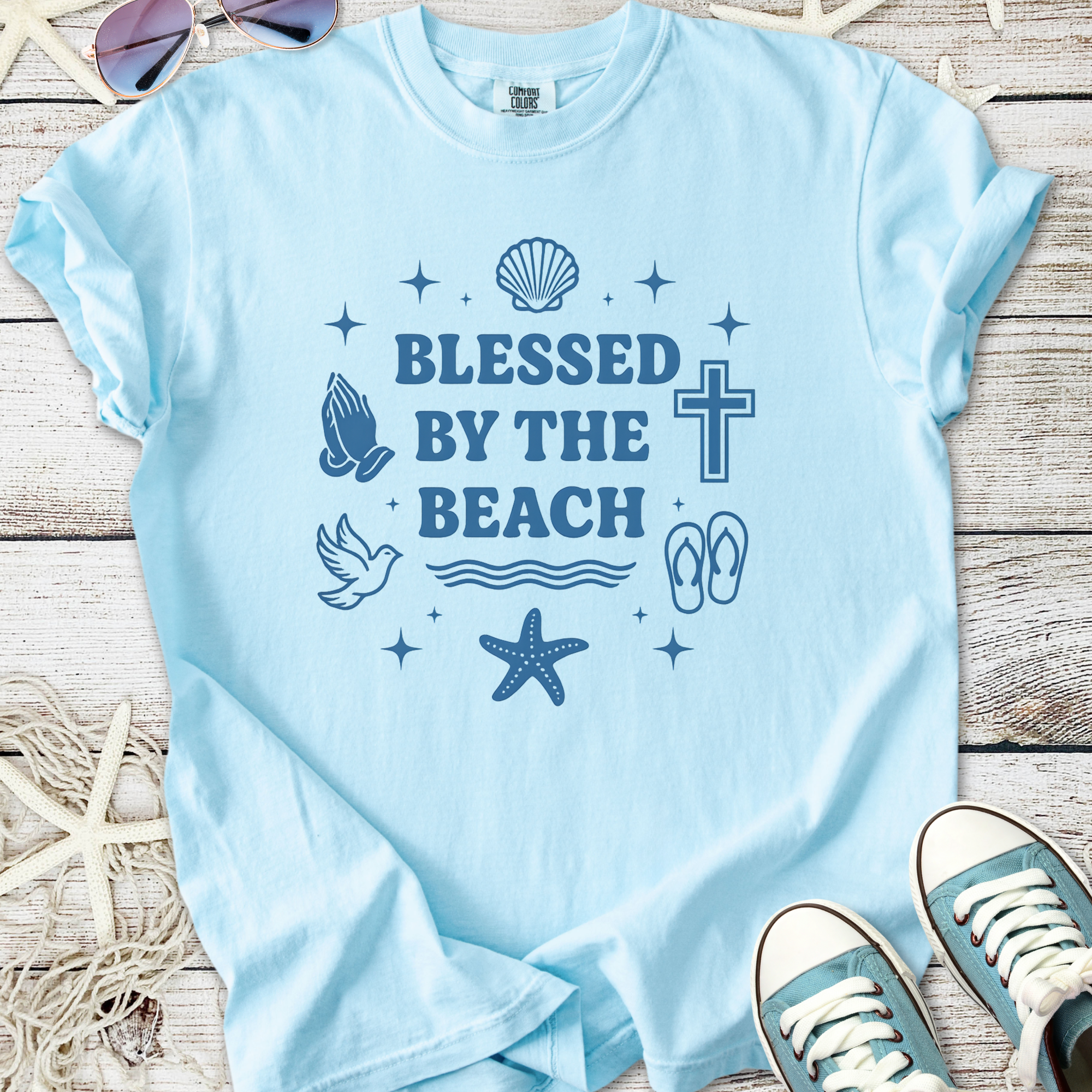 Light blue t-shirt with 'Blessed by the Beach' graphic featuring seashells, a cross, and beach motifs.