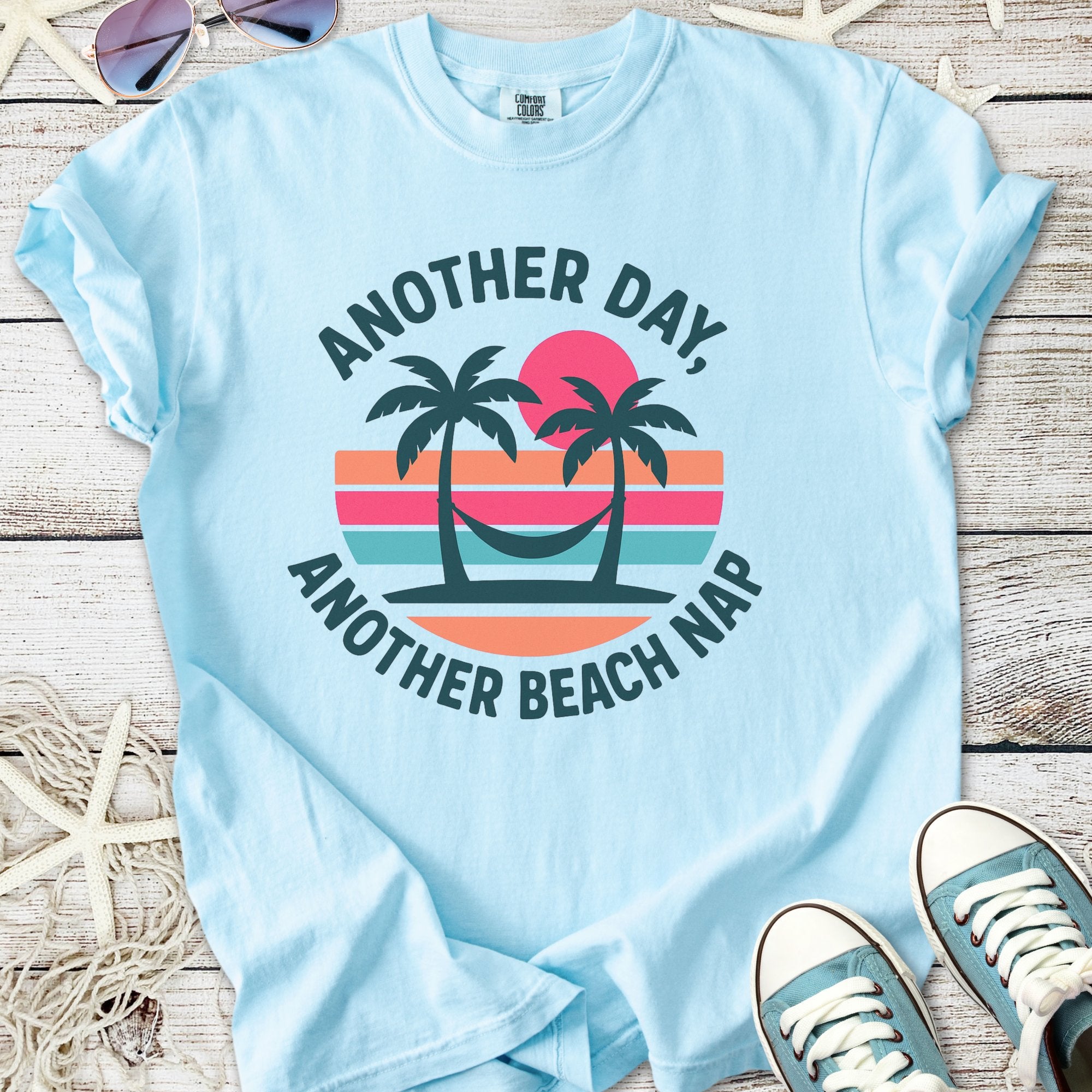 Light blue t-shirt with 'Another Day, Another Beach Nap' design featuring palm trees and sunset graphics.