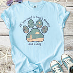 Light blue t-shirt with a beach-themed graphic: paw print design featuring waves, sun, and dog.