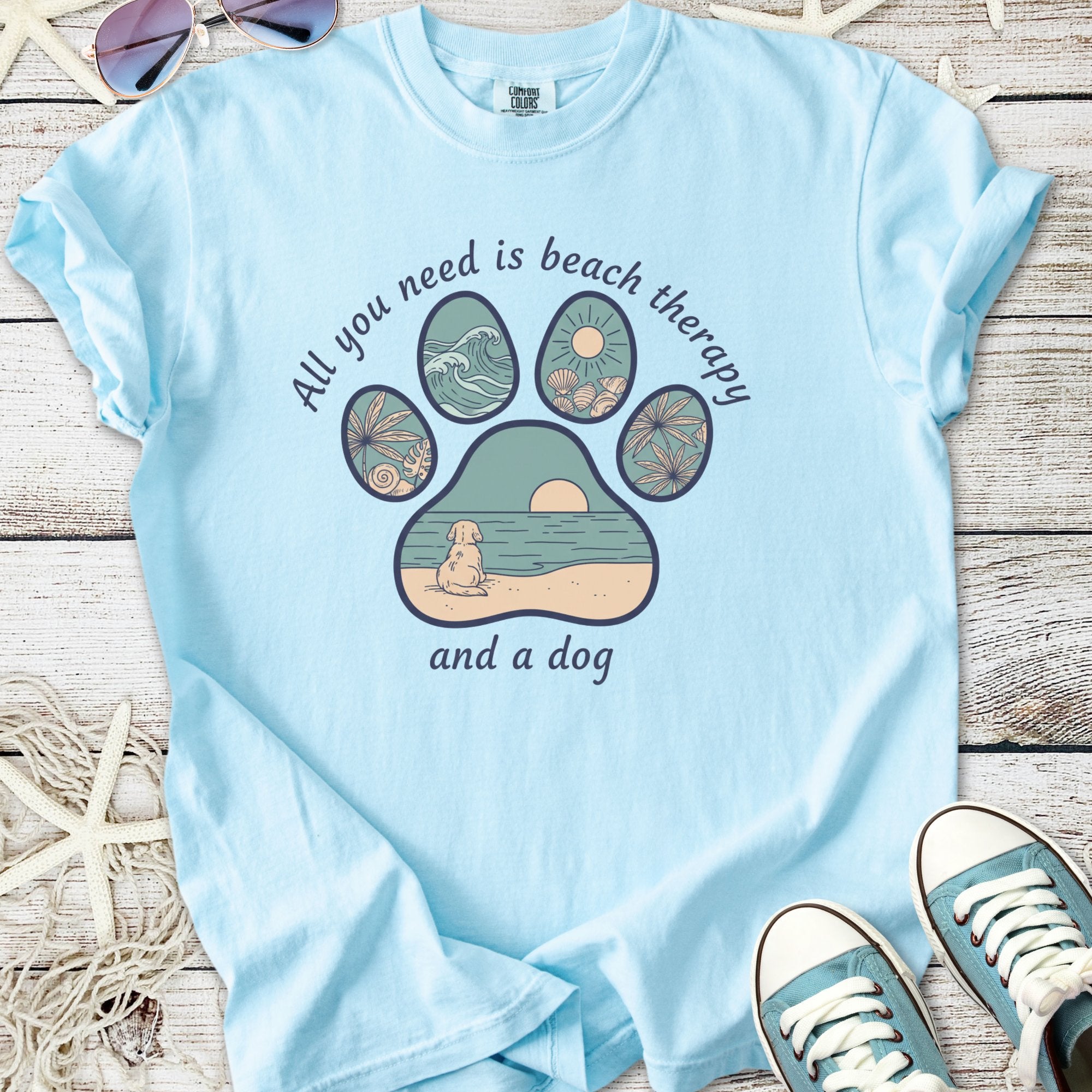 Light blue t-shirt with a beach-themed graphic: paw print design featuring waves, sun, and dog.