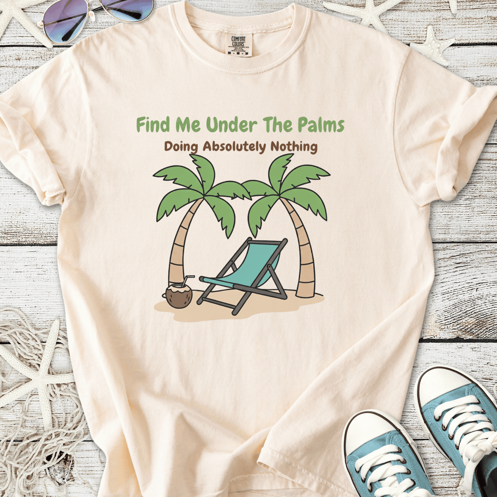 Light peach t-shirt with 'Find Me Under The Palms' graphic and a beach scene featuring palm trees and a lounge chair.