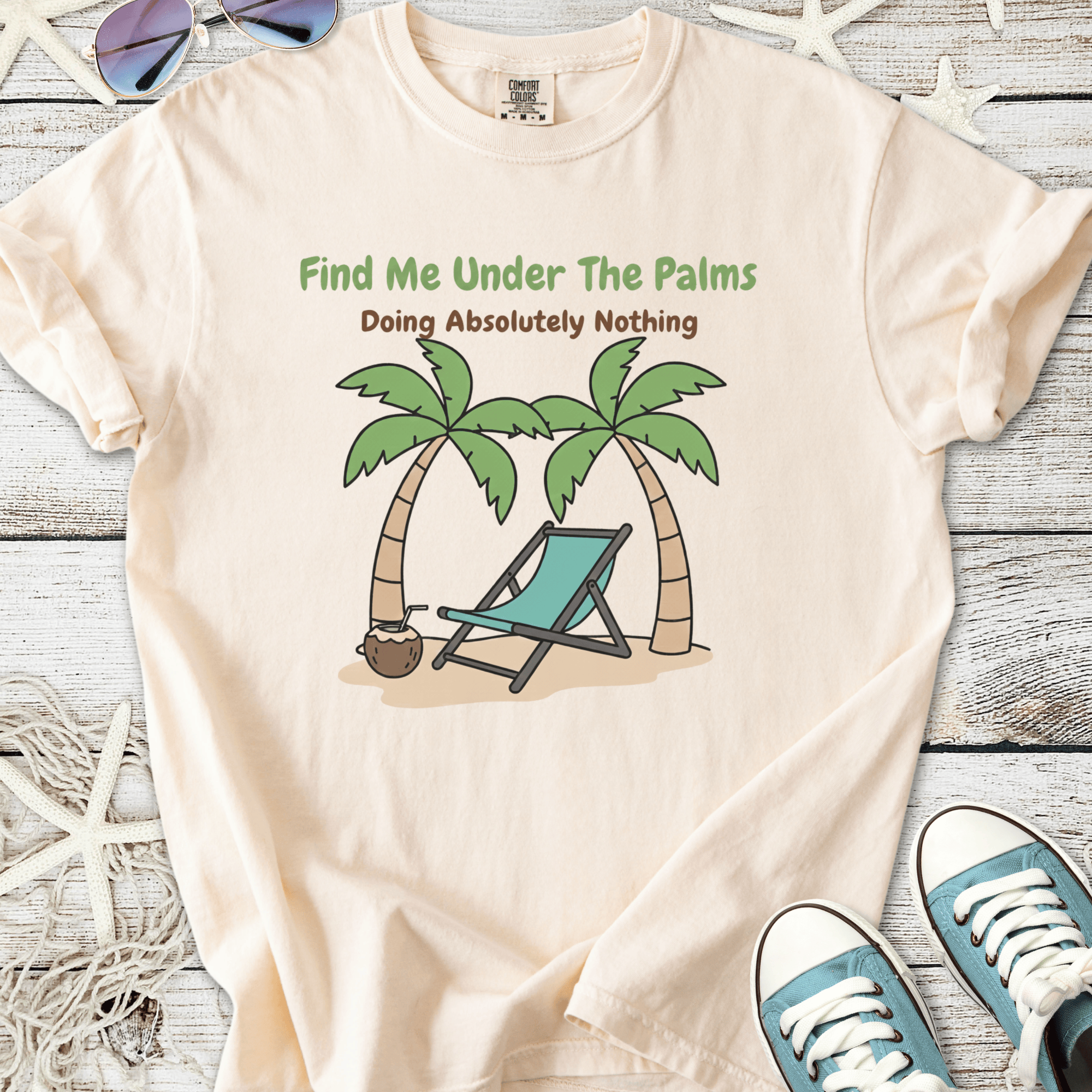 Light peach t-shirt with 'Find Me Under The Palms' graphic and a beach scene featuring palm trees and a lounge chair.