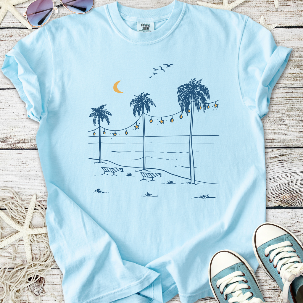Light blue t-shirt featuring a beach scene with palm trees, moon, and string lights, perfect for summer.