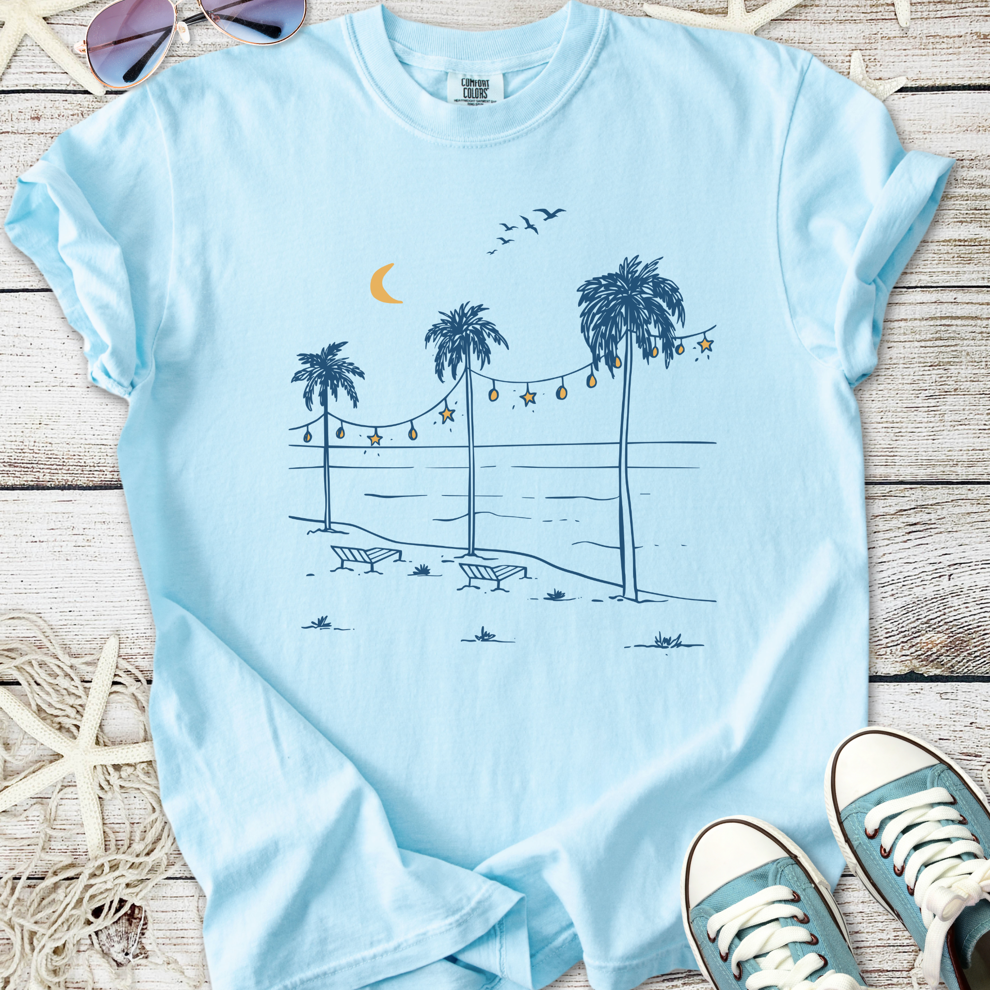 Light blue t-shirt featuring a beach scene with palm trees, moon, and string lights, perfect for summer.