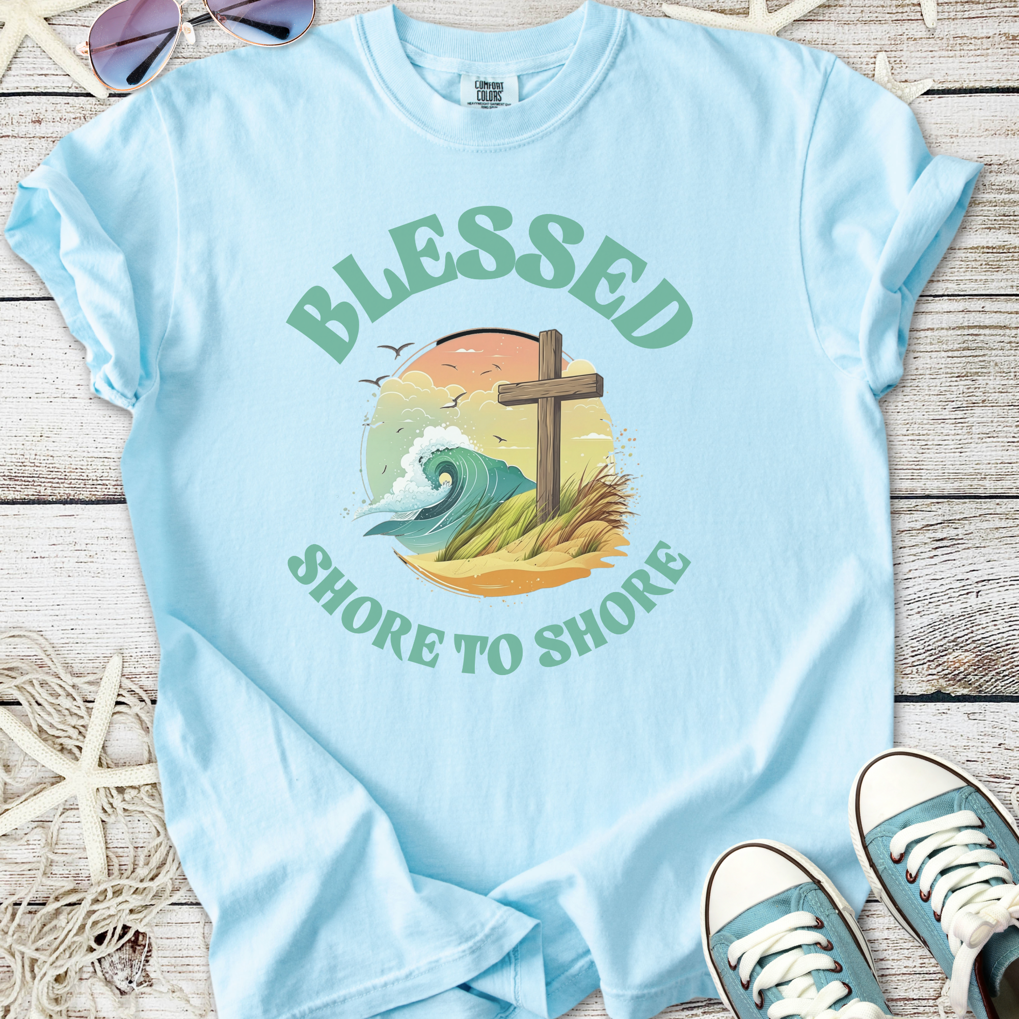 Light blue t-shirt featuring 'Blessed Shore to Shore' with wave and cross illustration.