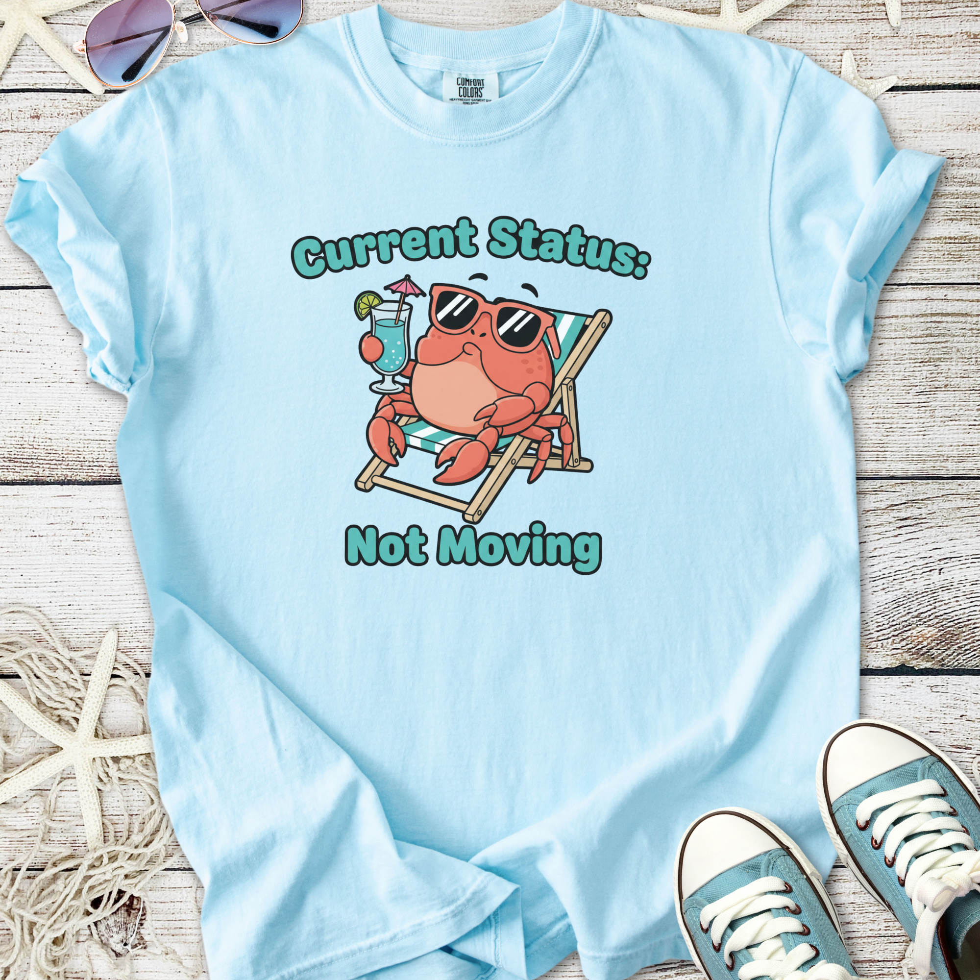 Light blue t-shirt featuring a playful crab graphic with sunglasses, lounge chair, and cocktail, ideal for summer.