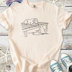 Cream graphic t-shirt featuring a playful dog on a dock with a fish, soft cotton material for comfort.