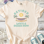 Light peach unisex t-shirt with 'Retired Life's a Beach Now' graphic and hammock design.