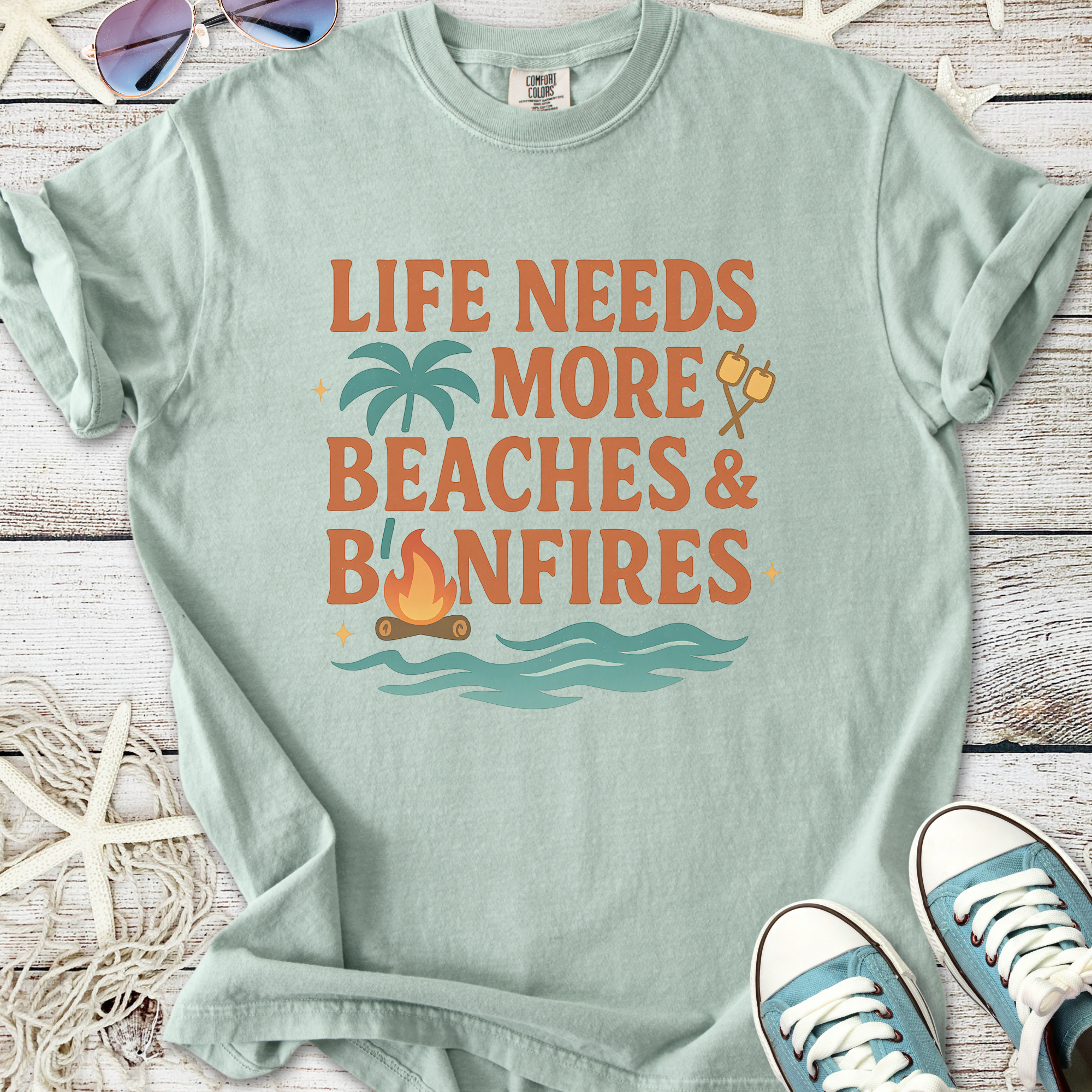 Light blue t-shirt with 'Life Needs More Beaches & Bonfires' graphic, perfect for beach lovers.