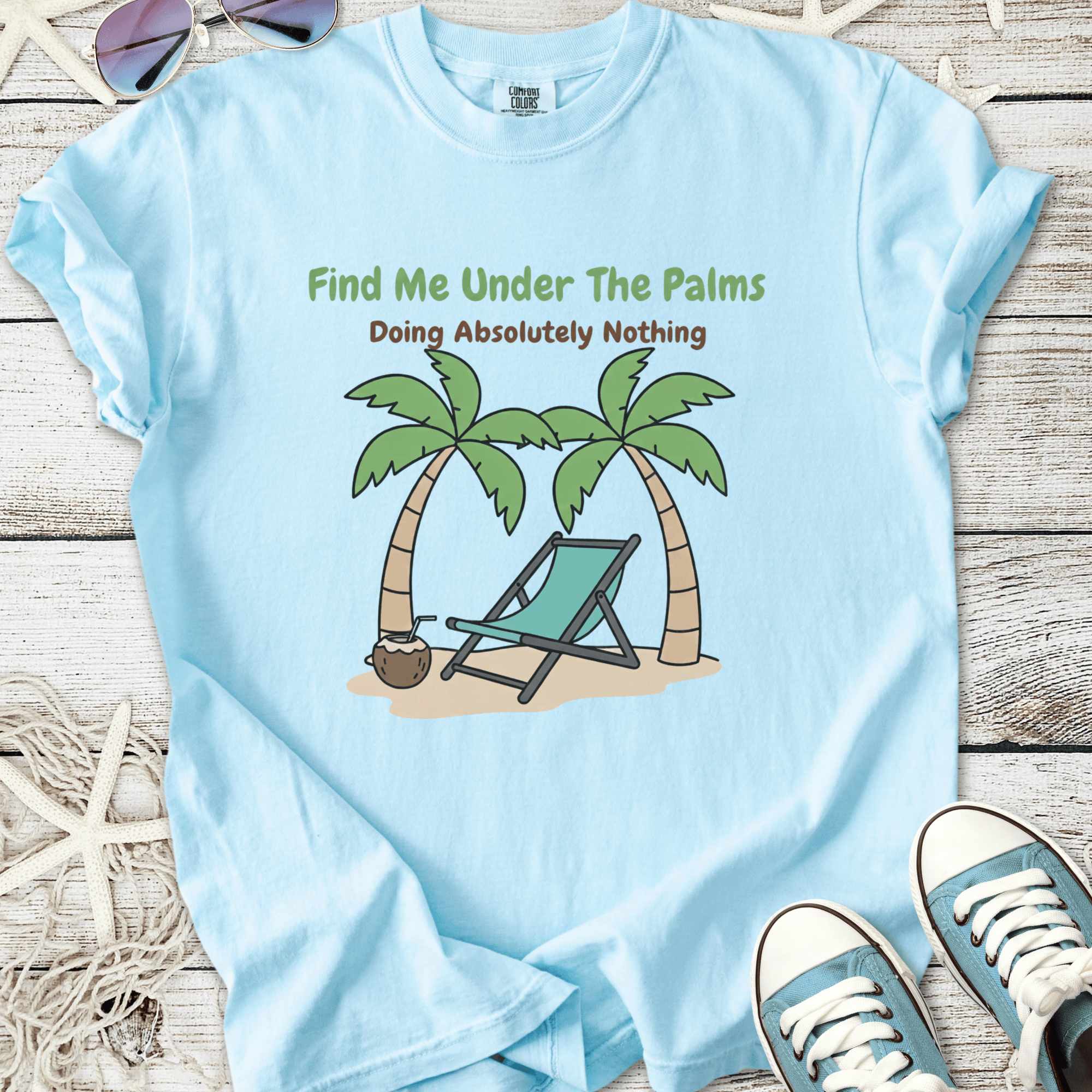 Light blue t-shirt featuring tropical palm trees and lounge chair graphic with the text 'Find Me Under The Palms'.