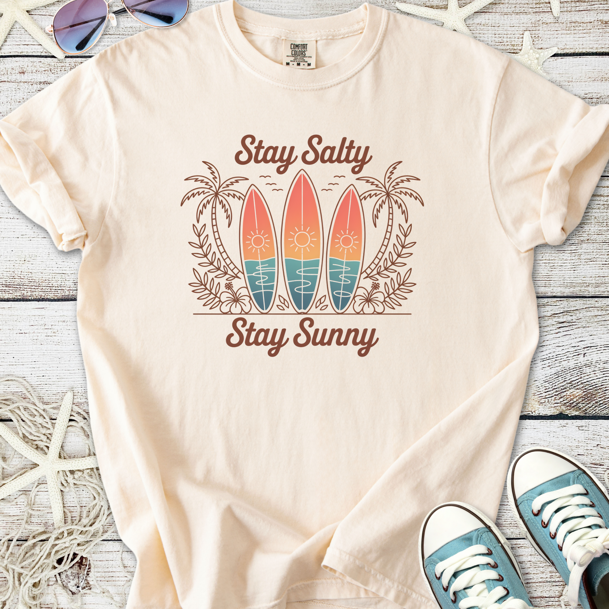 Beige graphic t-shirt featuring surfboards, palm trees, and the text 'Stay Salty Stay Sunny'