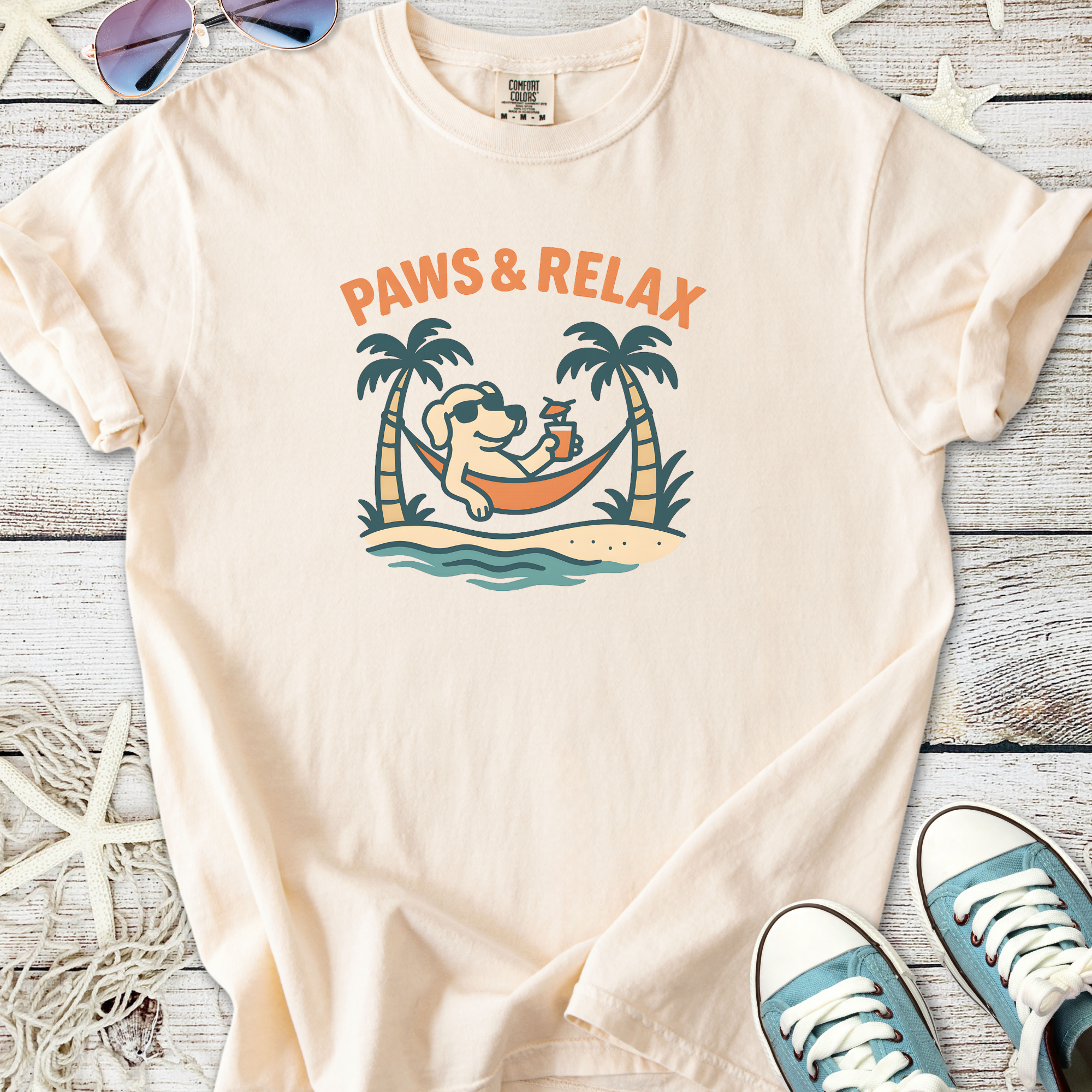 Light peach t-shirt with 'Paws & Relax' graphic featuring a dog on a hammock surrounded by palm trees.