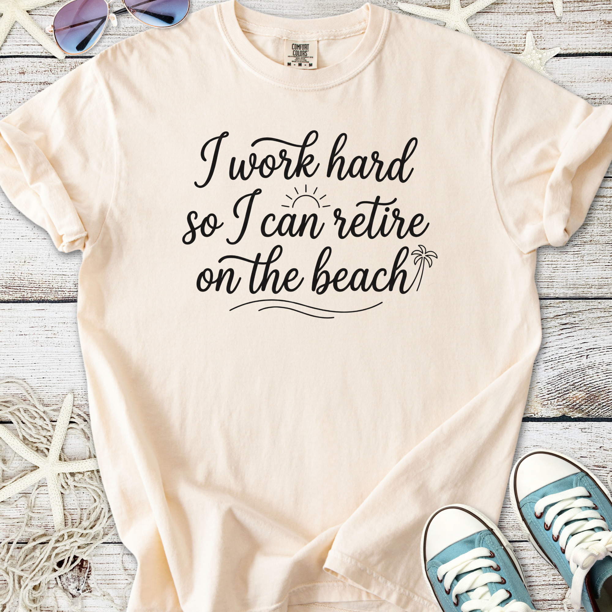 Light beige t-shirt with 'I work hard so I can retire on the beach' slogan, soft cotton material.
