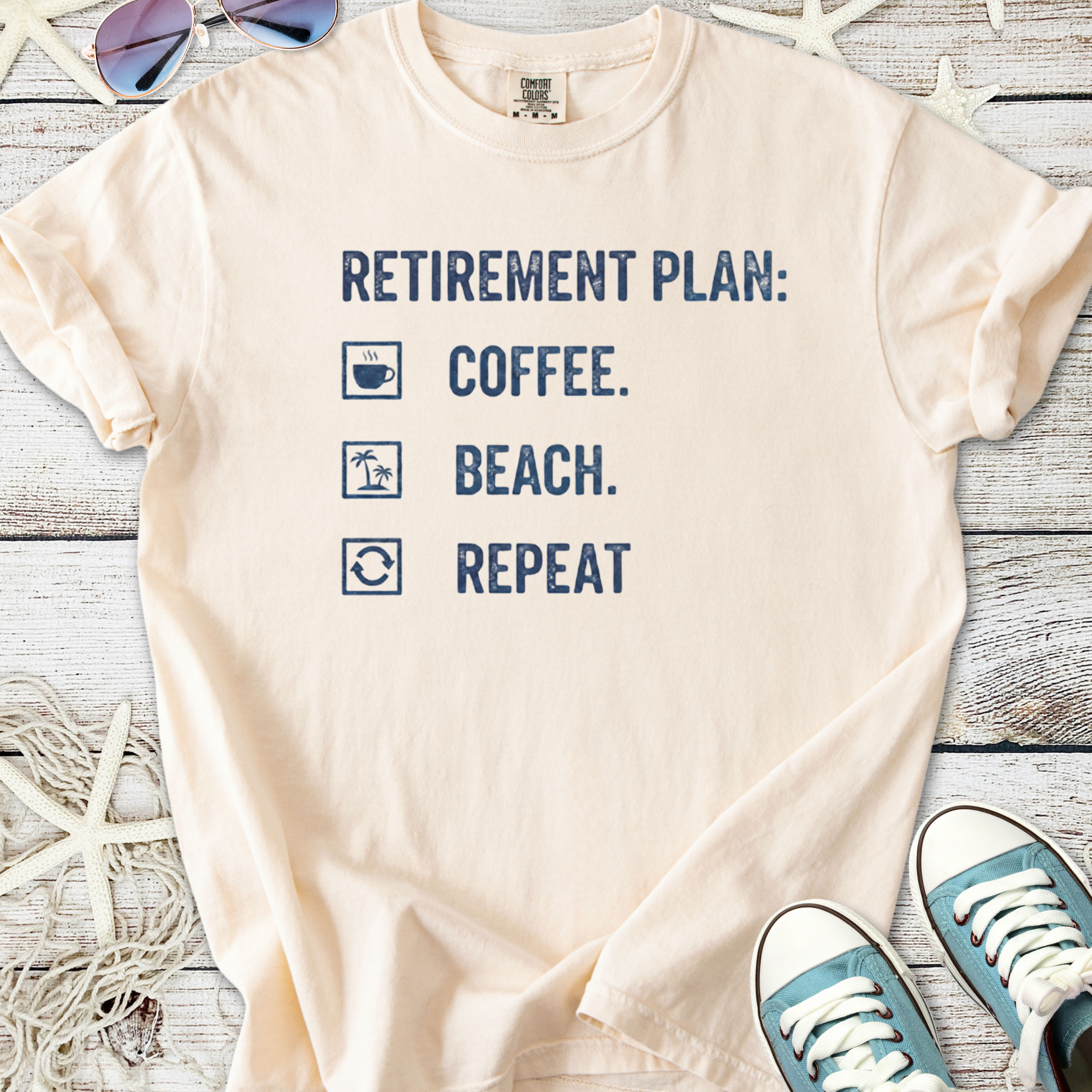 Light peach retirement plan t-shirt featuring coffee, beach, and repeat graphics in navy blue.