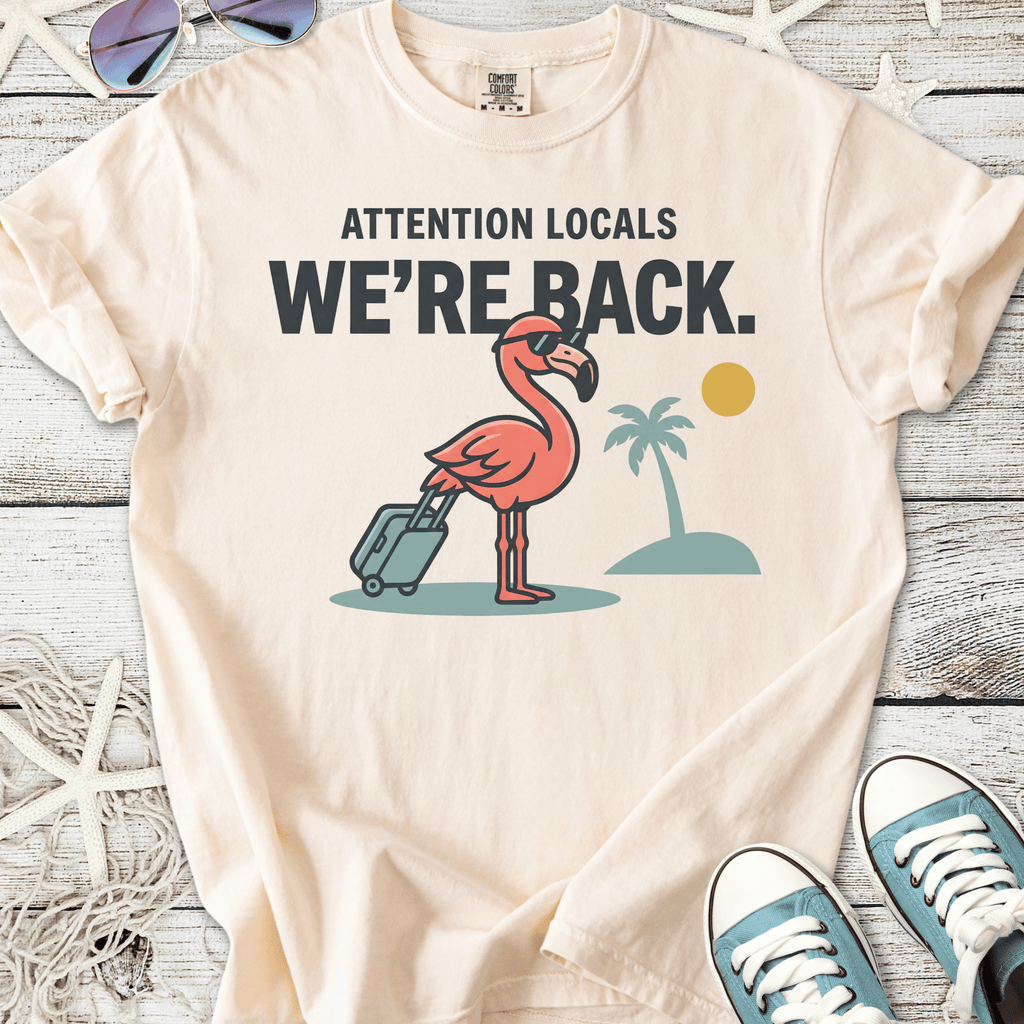 Beige t-shirt with a pink flamingo graphic and text 'Attention Locals, We're Back.' Perfect summer style.