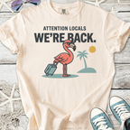 Beige t-shirt with a pink flamingo graphic and text 'Attention Locals, We're Back.' Perfect summer style.