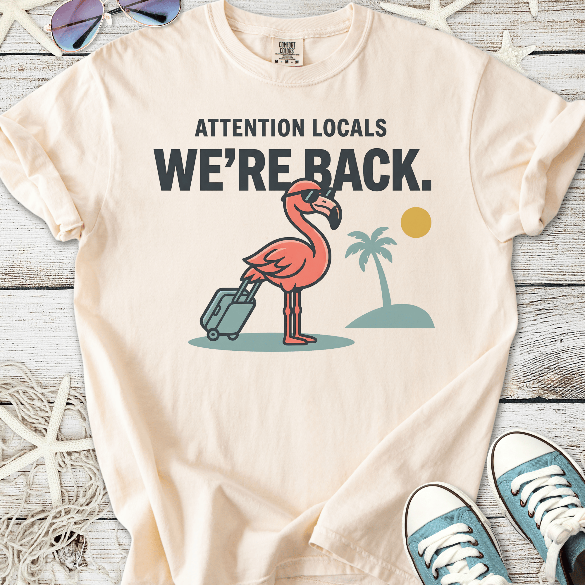 Beige t-shirt with a pink flamingo graphic and text 'Attention Locals, We're Back.' Perfect summer style.