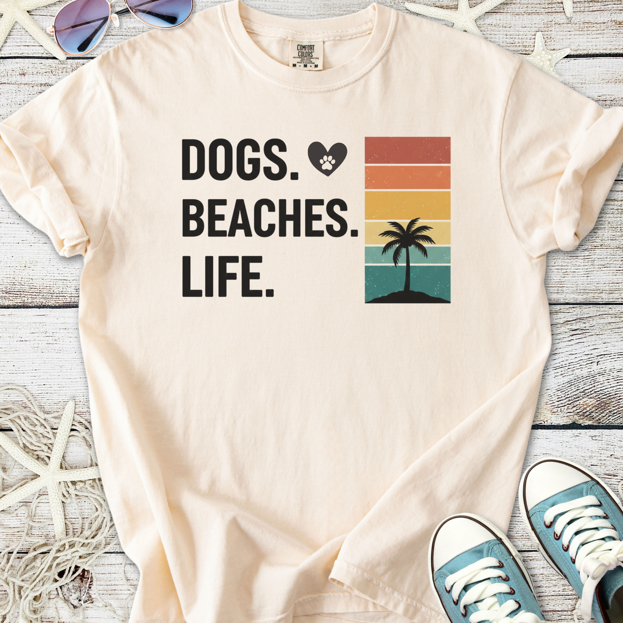 Cream t-shirt with 'Dogs. Beaches. Life.' graphic, featuring a palm tree and sunset colors, perfect for beach lovers.