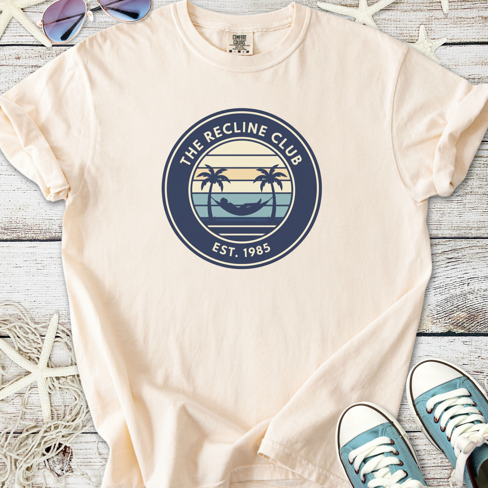 Beige cotton t-shirt featuring 'The Recline Club' logo with palm trees and hammock, est. 1985.