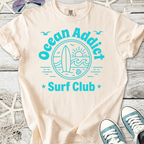 Beige cotton t-shirt with 'Ocean Addict Surf Club' graphic in teal and a surfboard illustration.