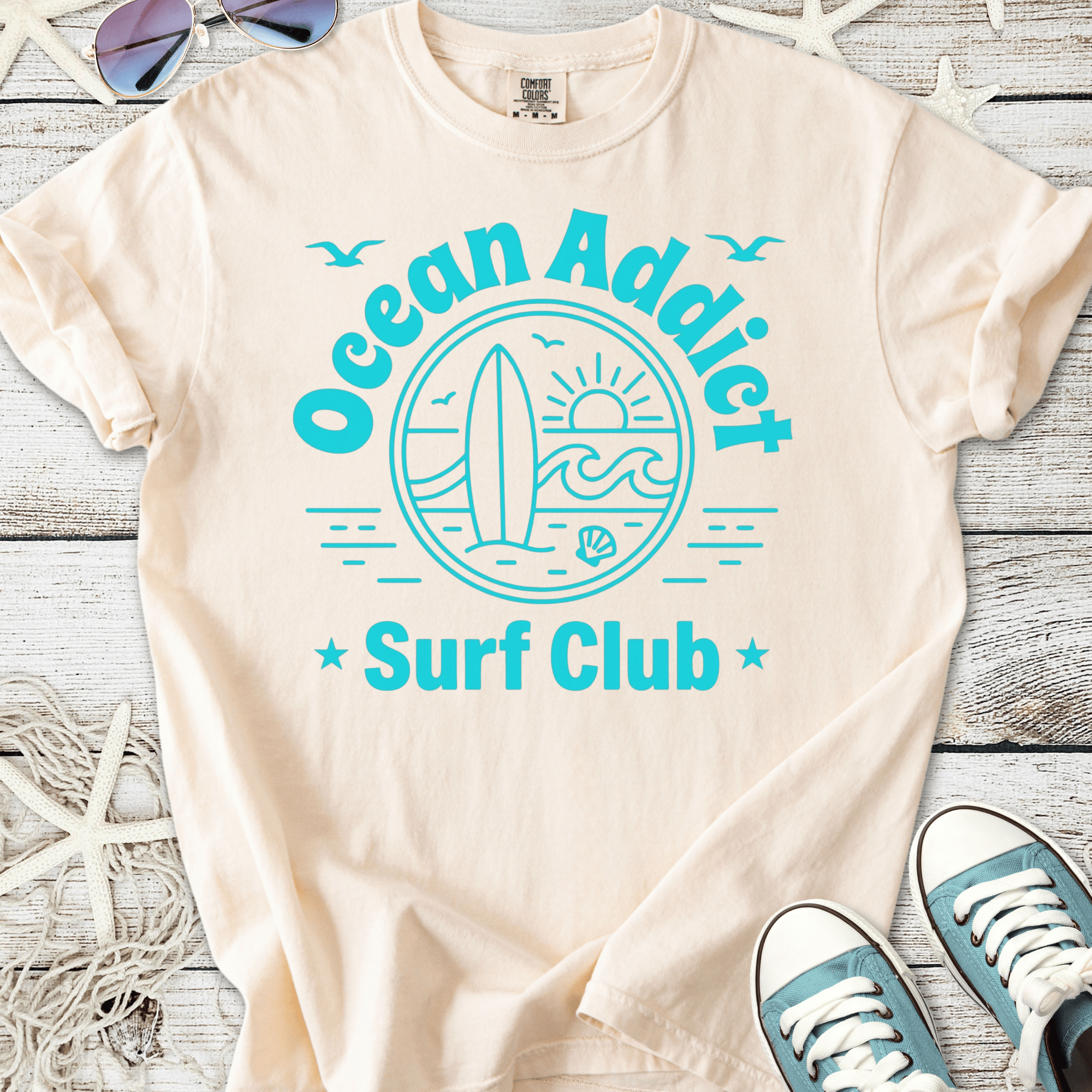 Beige cotton t-shirt with 'Ocean Addict Surf Club' graphic in teal and a surfboard illustration.