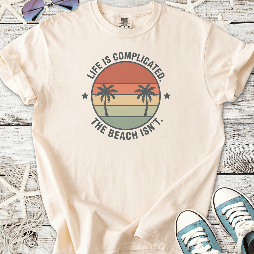 Light beige t-shirt with palm tree graphic and 'Life is Complicated. The Beach Isn’t.' slogan.