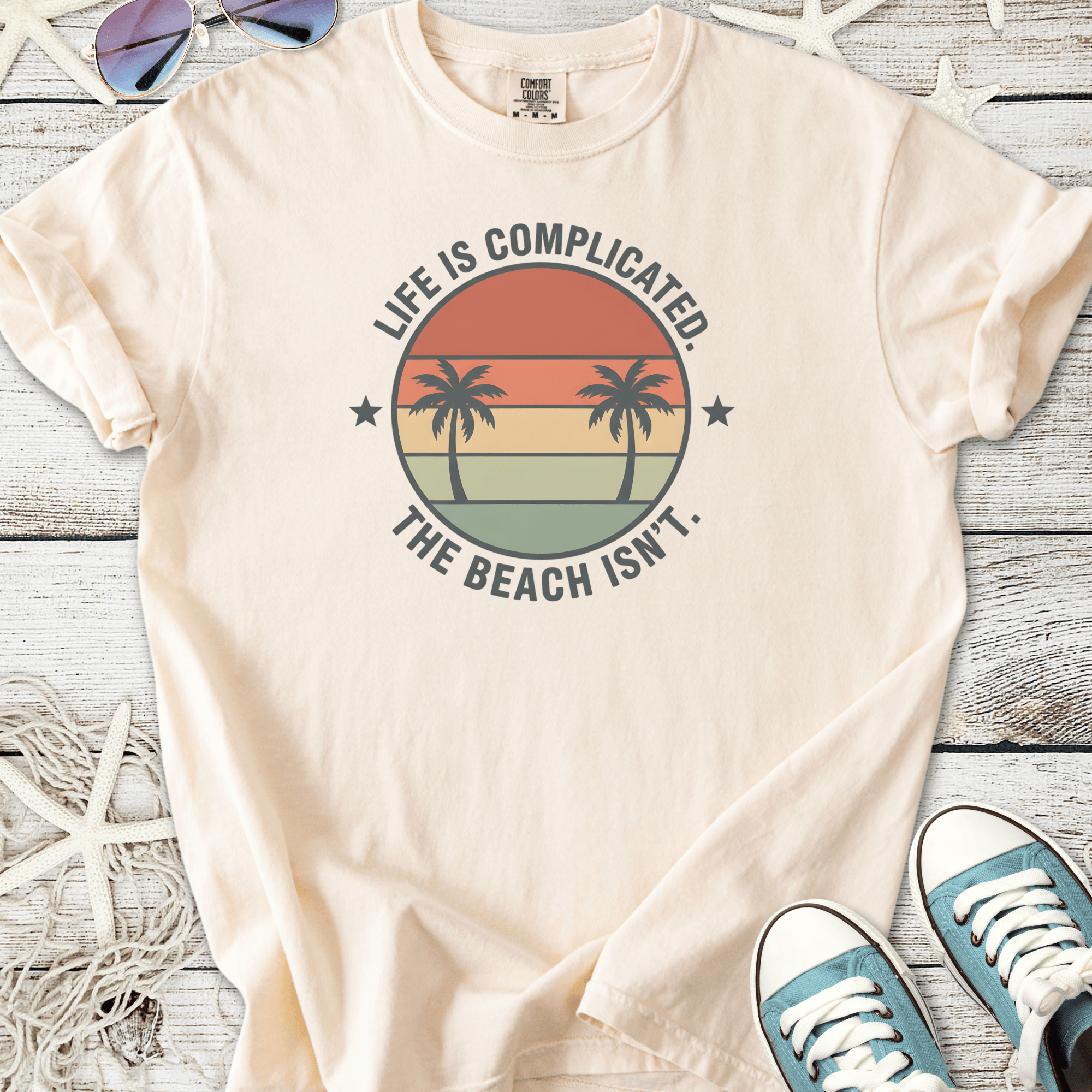 Light beige t-shirt with palm tree graphic and 'Life is Complicated. The Beach Isn’t.' slogan.