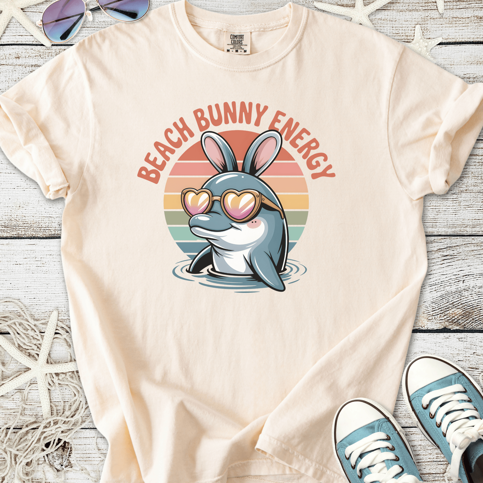 Cream t-shirt featuring a dolphin with bunny ears and heart-shaped sunglasses, text reads 'Beach Bunny Energy'.