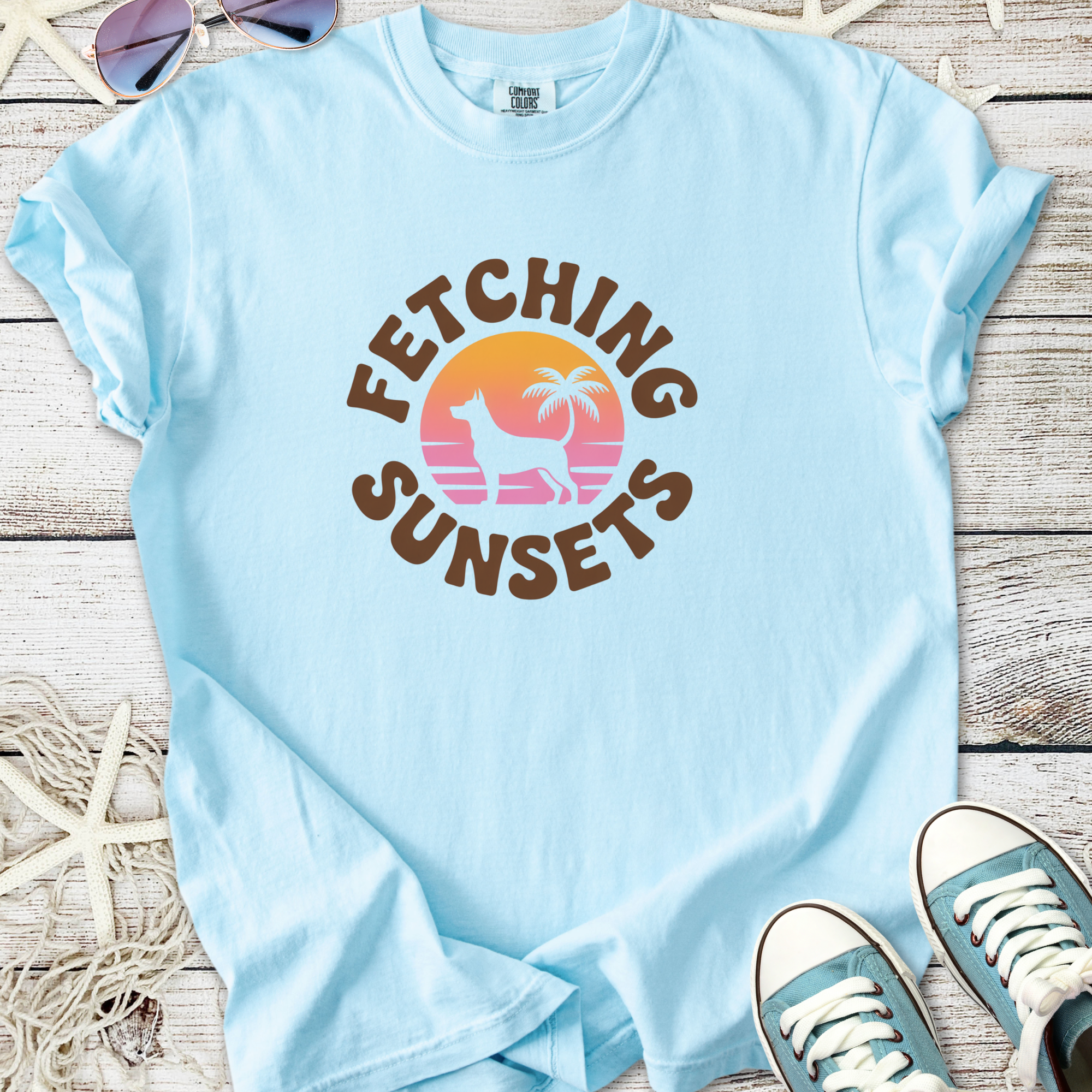 Light blue graphic tee 'Fetching Sunsets' with dog, palm tree design, perfect for casual summer wear.