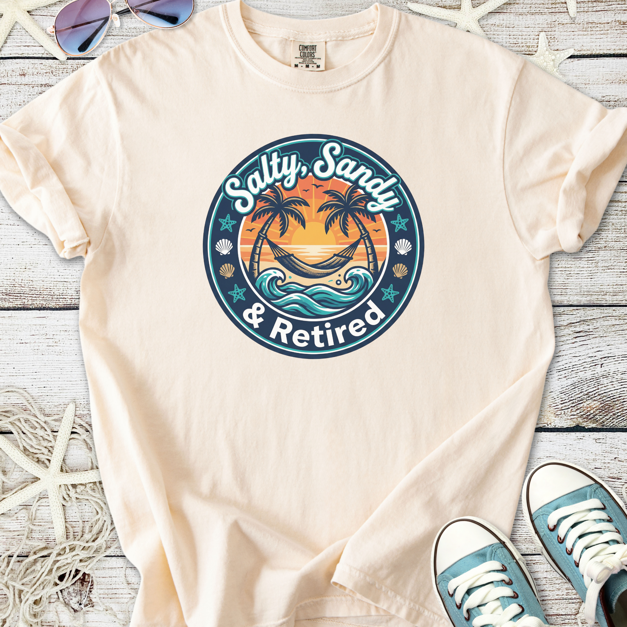 Beige t-shirt featuring 'Salty, Sandy & Retired' graphic with palm trees and sunset design.
