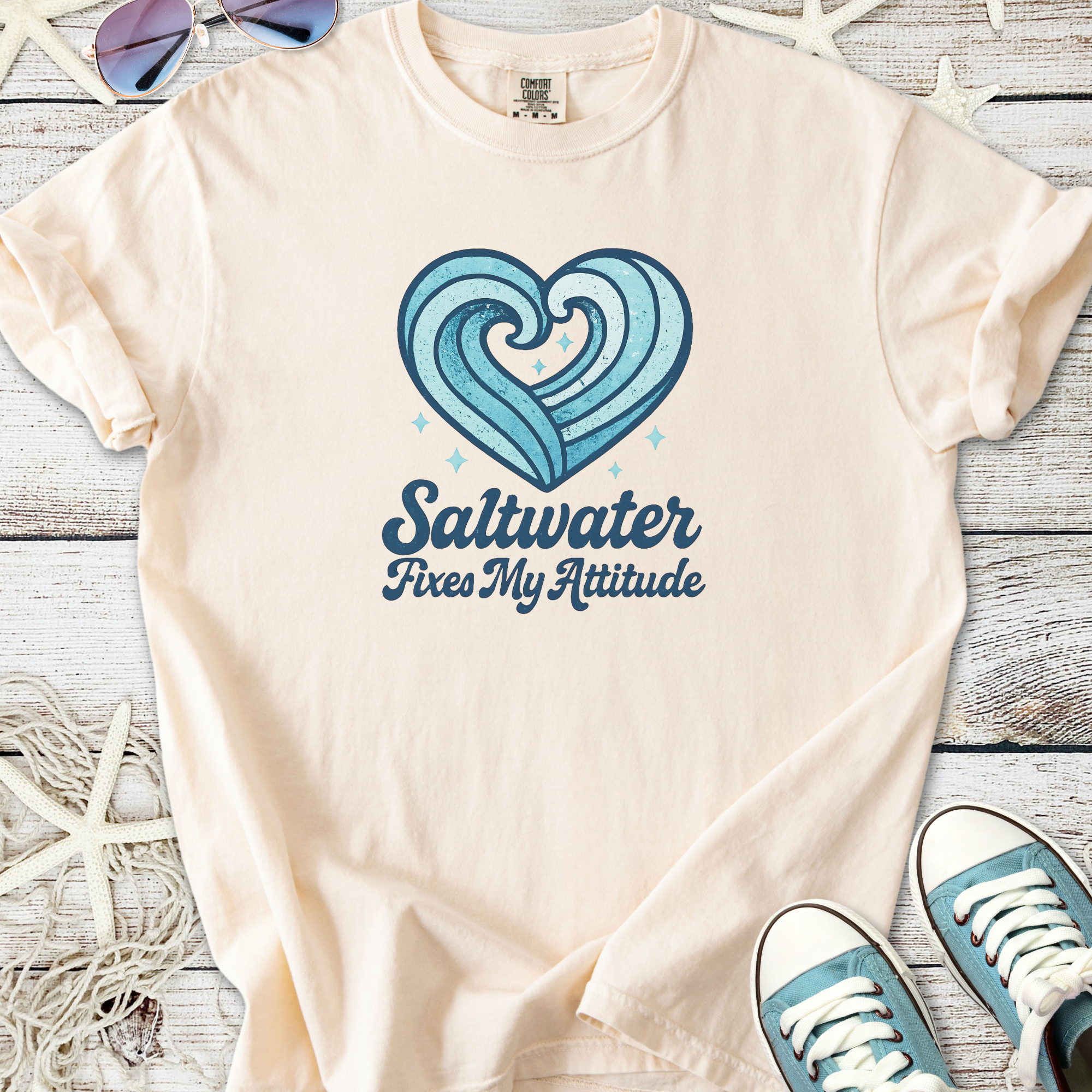 Beige graphic t-shirt with blue wave heart design and 'Saltwater Fixes My Attitude' text.