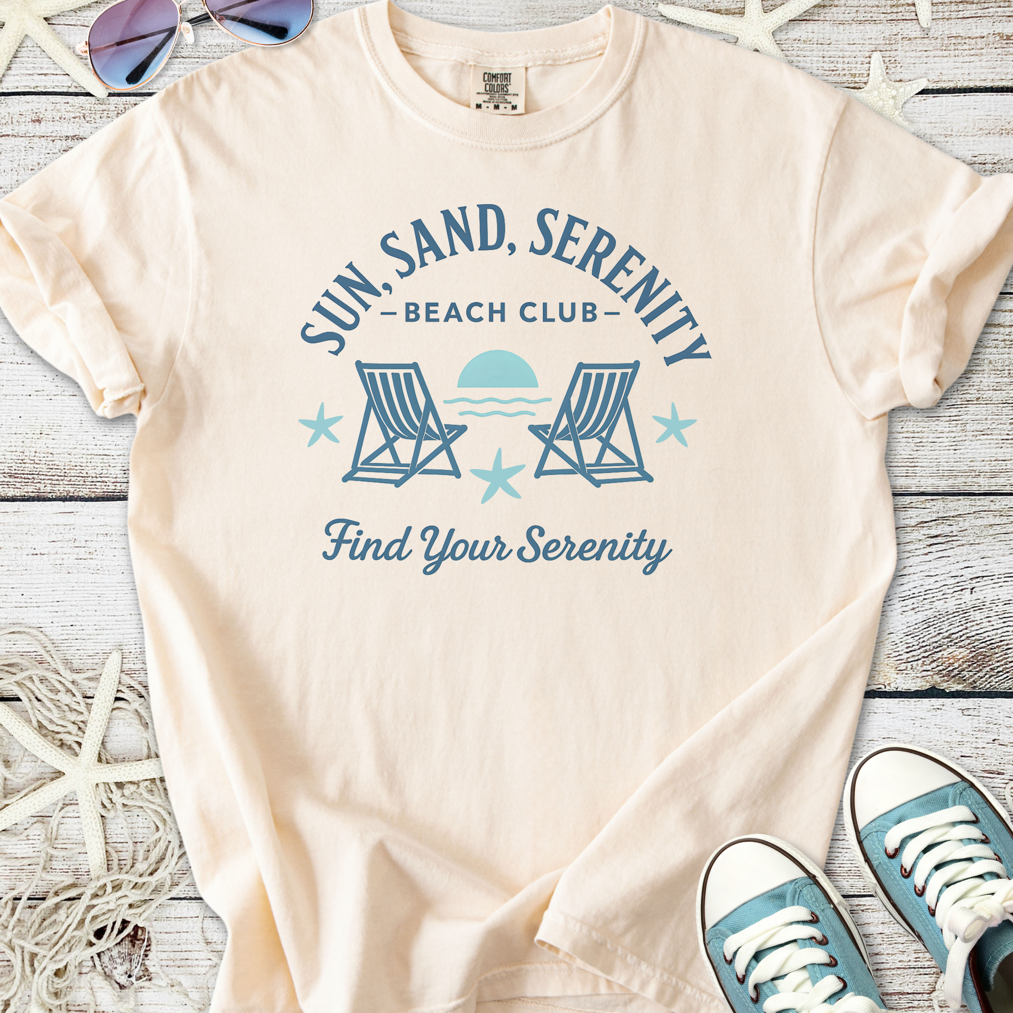 Beige cotton t-shirt with 'Sun, Sand, Serenity Beach Club' graphic and lounge chairs design.
