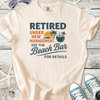 Light beige cotton t-shirt with ‘Retired Under New Management’ beach-themed graphic design.