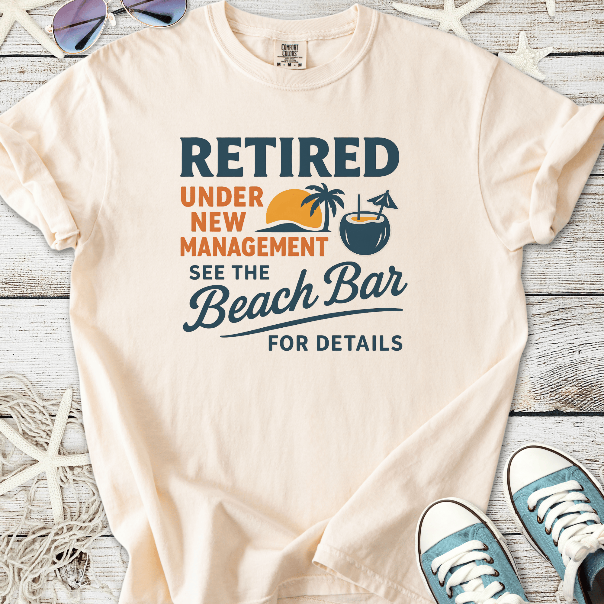 Light beige cotton t-shirt with ‘Retired Under New Management’ beach-themed graphic design.
