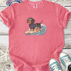 Pink t-shirt featuring a dachshund in sunglasses surfboarding, made of soft cotton.